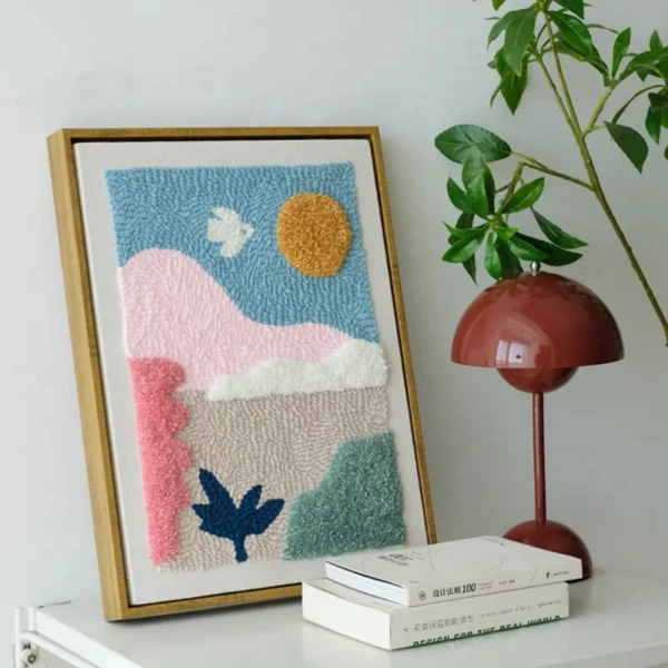Framed punch needle wall art featuring an abstract sunset valley scene with pink hills, blue sky, and sun, displayed on a shelf.