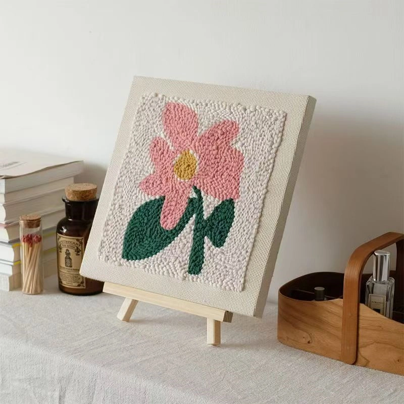 Punch needle flower embroidery on easel with pink bloom and green leaves, displayed in warm room
