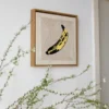 Andy Warhol banana punch needle art in wooden frame on wall with green botanical branches, artistic home styling