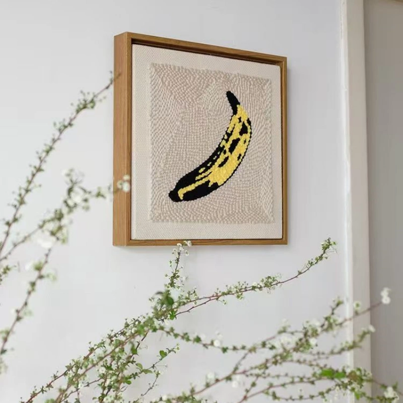 Andy Warhol banana punch needle art in wooden frame on wall with green botanical branches, artistic home styling