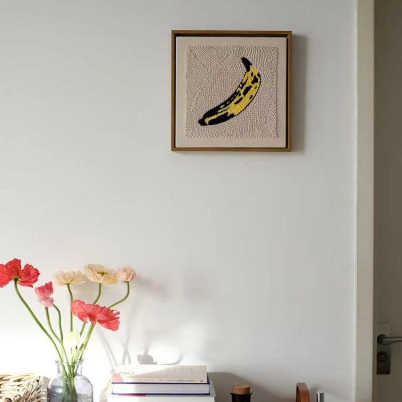 Framed Andy Warhol banana punch needle artwork hanging on white wall above flowers and books, modern interior design