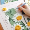 Hand using punch needle tool to create blooming daisy design with soft yarn on canvas.