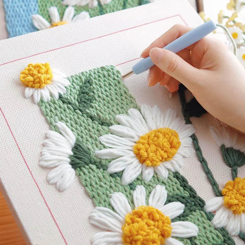 Hand using punch needle tool to create blooming daisy design with soft yarn on canvas.