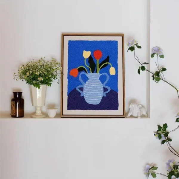Punch needle wall hanging art kit showing blue vase with red and yellow flowers on textured fabric, framed and displayed