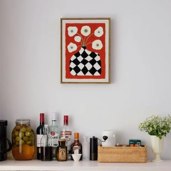 DIY wall decor punch needle art showing black checkered vase with white flowers on red fabric, framed and displayed above bar area