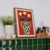 Framed punch needle wall art with a green vase and yellow flowers in bold colors on a red background.